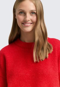 Czerwony sweter z dzianiny, z prążkowanym, okrągłym dekoltem i miękkim, teksturowanym wykończeniem. Modelka ma długie włosy, a tło jest neutralne.