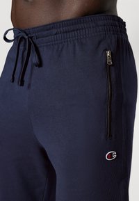 Marineblauwe joggingbroek met een elastische tailleband en verstelbaar trekkoord. Bevat een ritszak aan de zijkant en een klein logo-opdruk.