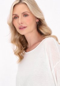 Fehér, könnyű kötött felső, széles nyakkivágással és laza fazonnal; puha textúrával és enyhe áttetszőséggel rendelkezik, amely kiemeli a természetes íveket.