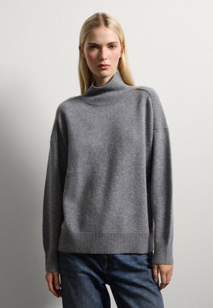 Maglione grigio chiaro a maglia con colletto alto, spalle cadenti e polsini a coste. Abbinato a jeans scuri e uno sfondo semplice.