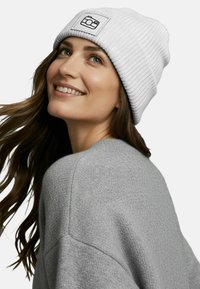 White ribbed knit beanie with a black camera logo patch. Model wears a soft gray sweater, showcasing the beanie's relaxed fit and texture.