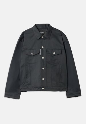 Veste noire surdimensionnée avec fermetures à pression, deux poches avant, portée sur une chemise grise. Un pantalon noir et des chaussures noires brillantes complètent le look.