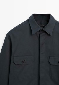 Dark grey button-up shirt made of cotton, featuring a pointed collar, two front flap pockets with buttons, and a smooth texture.