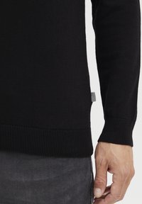 Svart strikket genser med ribbestrikkede mansjetter og nederkant, med et lite svart-og-hvitt merke på siden. Myk tekstur og figursydd design.