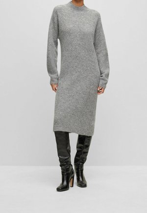 Persoon draagt een lang grijs gebreide midi-jurk met lange mouwen, gecombineerd met zwarte kniehoge laarzen met hak, staand tegen een effen achtergrond.