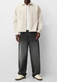 Giacca button-up beige chiaro sopra una maglietta bianca, abbinata a jeans larghi grigio scuro e sneakers color crema con una finitura testurizzata.