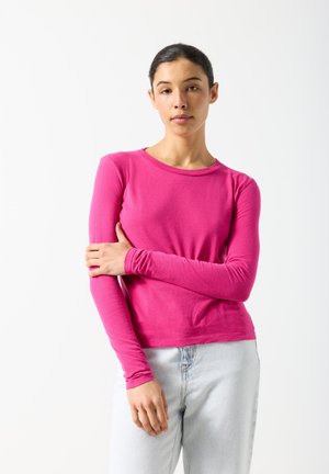 Mladá žena s tmavými vlasmi v ružovom tričku s dlhým rukávom a svetlomodrých džínsoch stojí na bielom pozadí, jej ľavá ruka sa dotýka pravej ruky.
