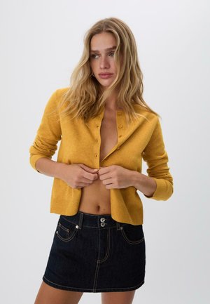 Jeune femme aux cheveux blonds lâches en train de boutonner un cardigan jaune moutarde, portant une mini-jupe en denim foncé, devant un fond clair et uni.
