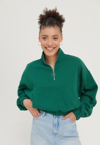 Žalia zip-up megztinis su aukštu apykakle, laisvu kirpimu ir susitraukusiais rankogaliais. Modelis derina jį su šviesiai mėlynomis aukštu juosmeniu džinsomis.