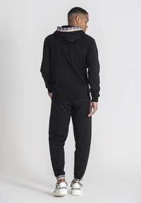 Svart hoodie med rutigt foder, parat med svarta joggingbyxor. Skorna har en vit sula med varumärke. Enkel, bekväm atletisk klädsel.