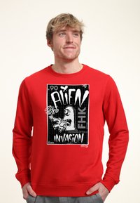 Červená mikina s kapucí s velkým černobílým grafickým motivem mimozemšťana a výrazným textem "ALIEN INVASION" na pozadí s mřížkou.