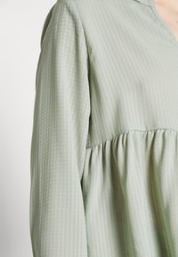 Blouse vert menthe avec un tissu texturé à rayures verticales, une coupe décontractée et des détails froncés à la taille.