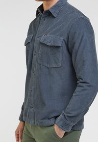Camicia in velluto a coste di colore blu scuro con righe verticali, chiusura frontale con bottoni, due tasche sul petto e colletto con bottoni. Texture morbida e design aderente.