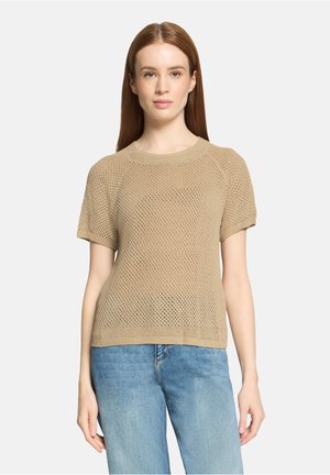 Woman with long red hair wearing a beige short-sleeve knit top and light blue jeans standing against a plain background.