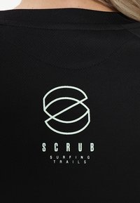 Schwarzes Sportshirt mit strukturiertem Stoff, einem runden Logo mit dem Text "SCRUB" über "SURFING TRAILS" in hellgrün.