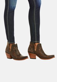 Botines de cuero marrón con punteras puntiagudas, costuras intrincadas y un tacón ligero, combinados con vaqueros ajustados azul oscuro.