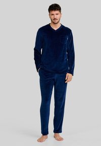 Hombre de pie descalzo, vistiendo una camiseta de manga larga y de canalé azul marino con escote en V, junto con pantalones a juego, sobre un fondo gris liso.