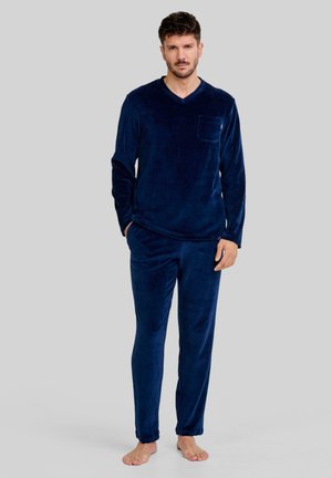 Hombre de pie descalzo, vistiendo una camiseta de manga larga y de canalé azul marino con escote en V, junto con pantalones a juego, sobre un fondo gris liso.