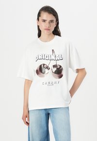 Balta per didelė medvilninė marškinė su grafika, vaizduojančia dvi vyšnias ir užrašu "ORIGINAL CHERRY CLUB" ryškiomis juodomis raidėmis.