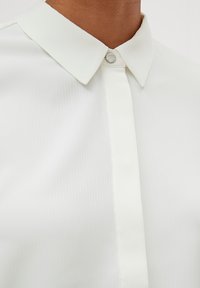 Chemise blanche à col en tissu côtelé, dotée d'une fermeture à boutons et d'un design épuré, avec une texture subtilement visible.