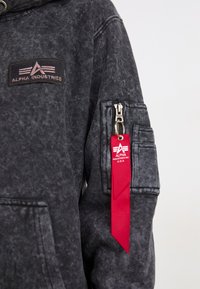 Schwarzer Denim-Hoodie mit strukturiertem Finish, einer Reißverschlusstasche am Ärmel und einem roten Stoffetikett mit dem Alpha Industries-Logo.