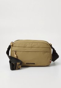 Compact beige crossbody bag with front zipper pocket, black adjustable strap, and small brand logo on front lower edge.