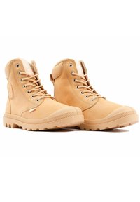 Botines tobilleros de color marrón claro, hechos de cuero con una superficie texturada, suela de goma, punta redonda y ojetes de metal para los cordones. Tacón reforzado.