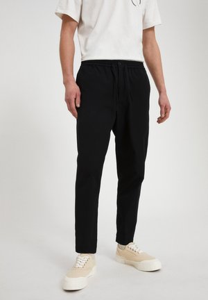 Uomo che indossa pantaloni neri con coulisse, t-shirt bianca e sneaker beige, in piedi davanti a uno sfondo chiaro e semplice.
