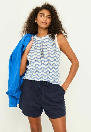 Femme aux cheveux bouclés portant un haut sans manches à motifs ondulés bleu et blanc, un short bleu marine, tenant une veste bleu vif sur l'épaule.