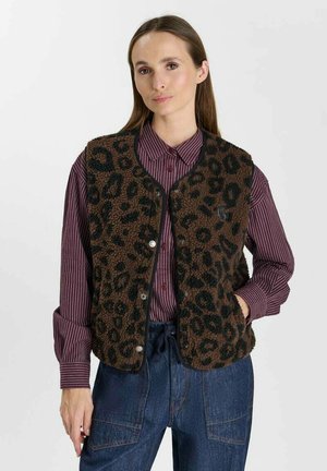 Vrouw draagt een bruine en zwarte fuzzy bodywarmer met luipaardprint over een paarse gestreepte blouse met knopen en blauwe spijkerbroek met handen in de zakken.