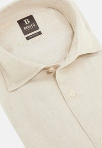Camicia di lino beige con vestibilità regolare, presenta una texture morbida, colletto standard, chiusura frontale con bottoni e un'etichetta nera con il nome del marchio.