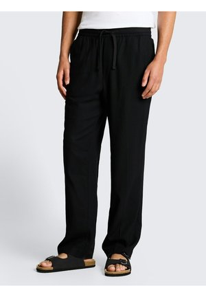 Mann trägt lockere schwarze Hose mit Kordelzug und schwarze offene Sandalen mit Korksohlen vor einem einfarbigen hellen Hintergrund.