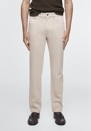 Mand iført beige bukser med lige ben, mørkebrune loafers og en mørkebrun striktrøje, stoppet ned i bukserne, stående med armene afslappet ned langs siderne.