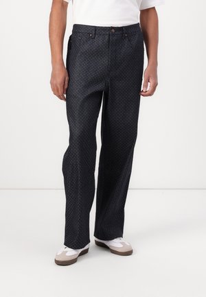 Dark denim trousers with a loose fit, featuring a polka dot pattern, button closure, and five pockets. Paired with white and beige sneakers.