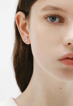 Mujer joven de piel clara y ojos azules, con un pequeño pendiente geométrico transparente en la oreja izquierda, sobre un fondo blanco.