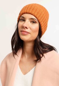 Beanie tricotat portocaliu cu textură în relief, material moale. Purtați cu un șal roz deschis peste un pulover alb cu decolteu în V.