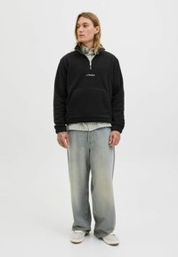 Person med langt hår iført en sort zip-up fleece fra JJ Studios over en ternet skjorte, løse falmede jeans og hvide sneakers, stående mod en hvid baggrund.