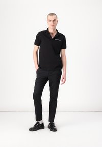 Polo nero con colletto a tre bottoni, logo ricamato sul lato sinistro del petto, abbinato a pantaloni neri slim e scarpe nere.