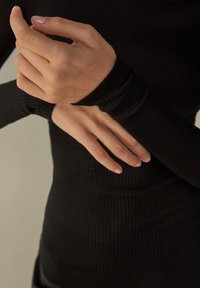 Černý žebrovaný top s dlouhými rukávy a přiléhavými manžetami. Detailní pohled na ruce, který zdůrazňuje strukturovanou tkaninu a detaily rukávu.