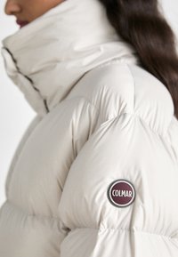 Bela puhovka z visokim ovratnikom, z okroglim burgundskim logotipom Colmar na rokavu in quiltnim teksturo.