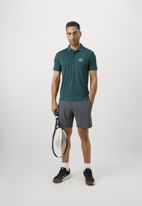 Vyras vilkintis mėlyną polo marškinėlį, pilkas šortus ir juodus sportinius batelius, laikantis teniso raketę, stovintis prieš paprastą šviesų foną.