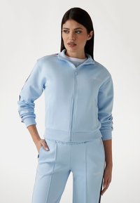 Lichtblauwe ritsjack van gladde stof, met een hoge kraag, zijzakken en geborduurd logo. Bijpassende broek zichtbaar.