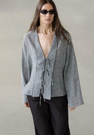 Femme portant des lunettes de soleil noires et une veste grise ample à manches longues avec des liens à l'avant, associée à un pantalon foncé, sur un fond uni.