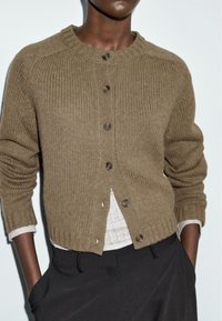 Bruine gebreide cardigan met geribbelde textuur, ronde halslijn, sluiting met knopen aan de voorkant en omgeslagen manchetten, gedragen over een lichtgekleurde top.