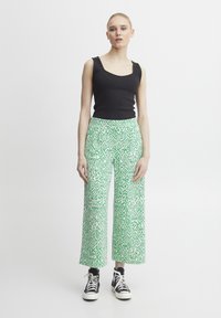 Débardeur noir côtelé associé à un large pantalon vert à motifs avec un design géométrique. Le modèle porte des baskets noires.