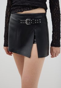 Mini falda de cuero negro con una abertura frontal y cinturón de hebilla plateada. Textura suave, diseño ajustado, combinada con una delicada blusa de encaje de manga larga.