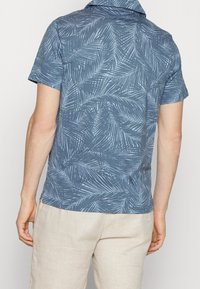 Chemise à manches courtes bleue avec un motif de feuilles blanches, tissu doux, détail de col et coupe décontractée, associée à un pantalon clair.