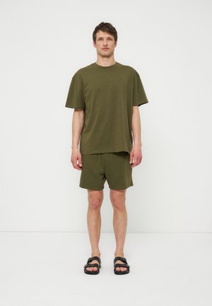 Άνδρας στέκεται σε λευκό φόντο φορώντας ελιά πράσινο κοντομάνικο T-shirt, ταιριαστά σορτς και μαύρα σανδάλια με αγκράφα.