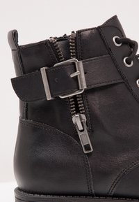 Bota negra de cuero hasta el tobillo con cremallera lateral, detalle de hebilla de metal, costuras en contraste y ojales redondos para los cordones.