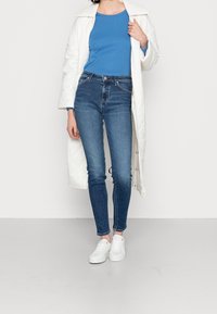 Witte gewatteerde lange jas, blauwe lange mouwen top en donkerblauwe hoog-getailleerde skinny jeans. Witte sneakers met een textuurafwerking.